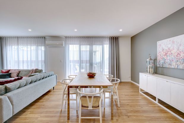 Bright open-plan dining and living area with a light timber floor, pale upholstered sectional sofa, white dining table and sculptural dining chairs. Floor-to-ceiling sheer curtains and glazed doors bring in abundant daylight, while a grey feature wall supports a white sideboard and soft pink abstract artwork.