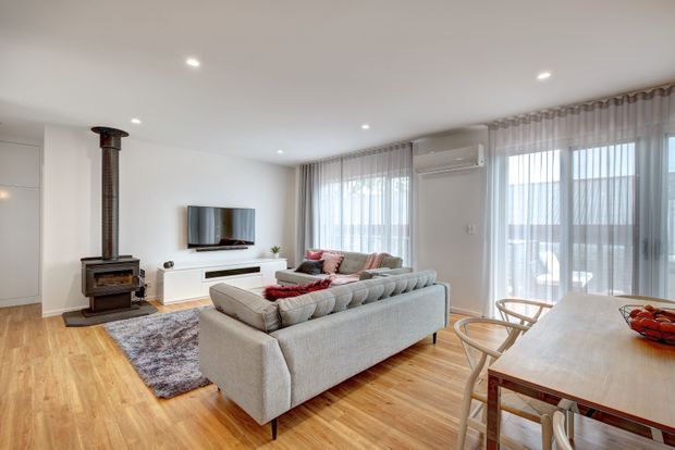 Bright open-plan living and dining area with warm timber flooring, a large light-grey sectional sofa, wall-mounted television and low white media cabinetry. A freestanding wood-burning stove sits against the left wall, while sheer floor-to-ceiling curtains and glazed doors bring in abundant natural light. The dining table occupies the foreground near the windows.