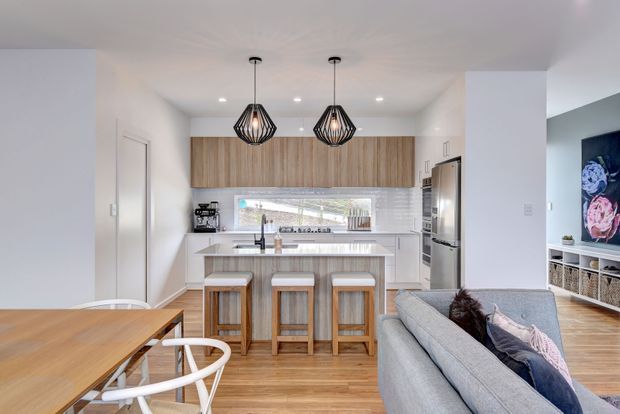 Bright open-plan kitchen and living area with a white island featuring seating for three, timber cabinetry, a tiled backsplash, stainless-steel appliances, timber flooring, and contemporary pendant lights. A dining table and sofa are visible in the adjoining living space.