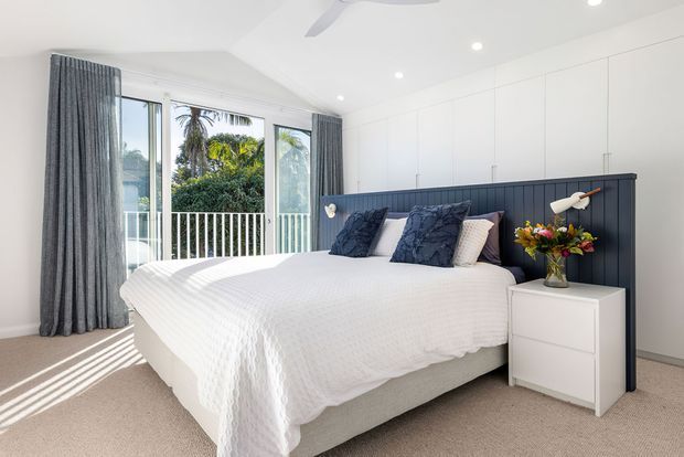 Bright contemporary bedroom with a large upholstered bed dressed in white bedding, a dark navy vertical-panel headboard, white built-in wardrobes, bedside furniture, recessed ceiling lights, and full-height glazed doors opening to a balcony with tropical greenery beyond. Neutral carpet and soft grey curtains create a clean, airy atmosphere.