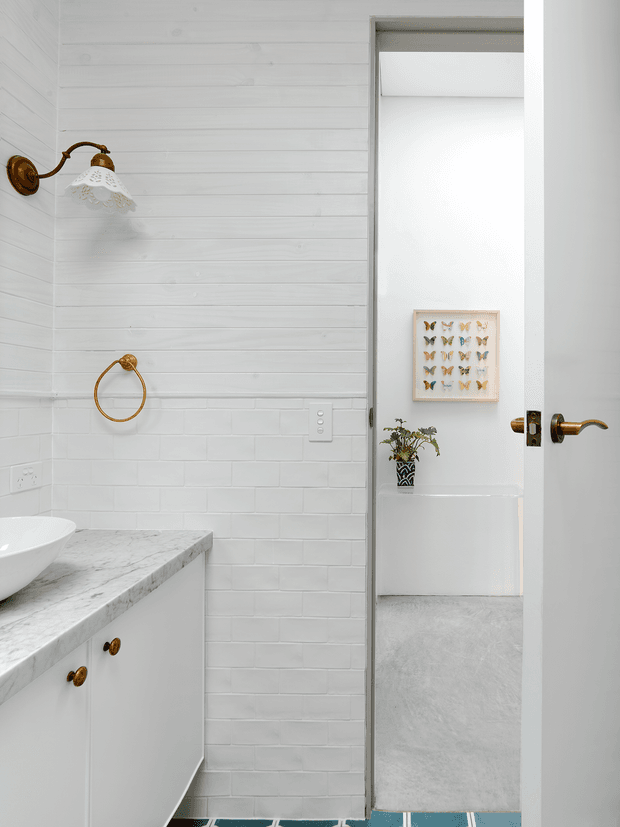 Bright white bathroom with horizontal white wall panelling above white subway tile, a white vanity with marble-look benchtop and vessel basin, and brass wall sconce, towel ring, cabinet knobs, and door hardware. An open door reveals an adjacent carpeted room with butterfly artwork and a small plant.