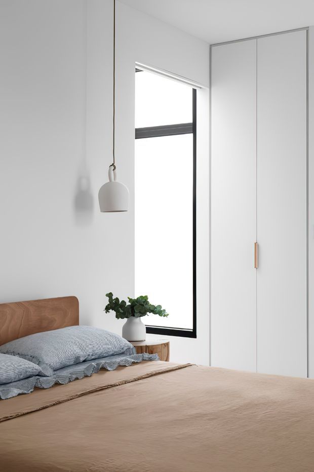 Bright, minimalist bedroom with a low timber bedhead, neutral beige bedding, textured blue-grey pillows, a small timber bedside table, white pendant light, potted plant, tall black-framed window, and full-height white built-in wardrobes with timber handles.