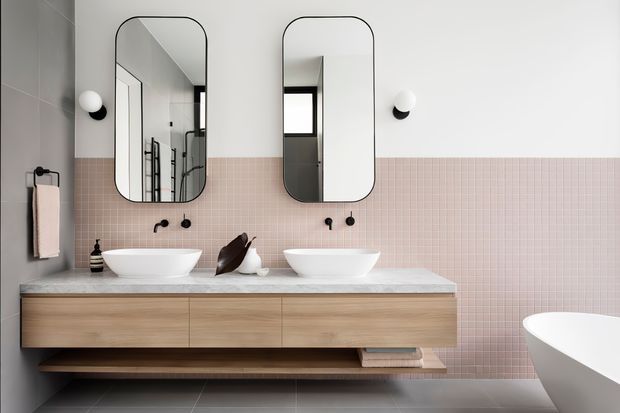 Contemporary double-vanity bathroom with a floating light timber wall-mounted vanity, two white vessel basins, pale pink small-format tiled splashback, large rounded-rectangle mirrors, black tapware and wall lights, and a freestanding white bathtub partially visible at right. The space has a clean, refined, softly warm palette with dark grey floor and wall tiles.