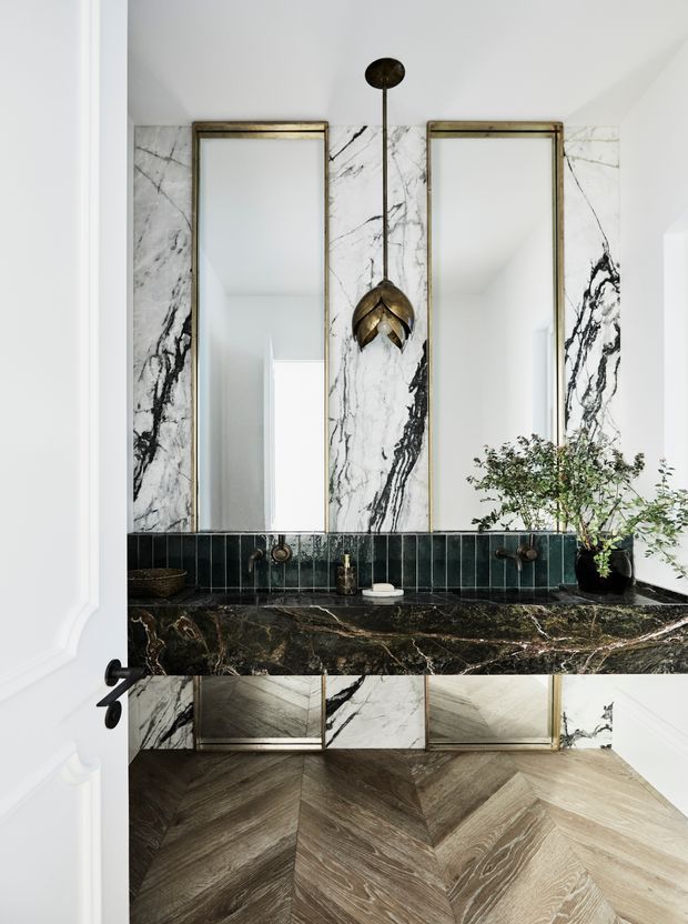 A refined bathroom vanity viewed through an open doorway, featuring a dramatic white-and-black veined marble wall, dark marble benchtop, deep green vertical tile splashback, twin aged-metal framed mirrors, wall-mounted bronze-toned tapware, and a sculptural pendant light. The room has a warm chevron-patterned timber floor and white decorative wall panelling.