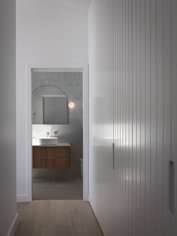 A narrow, softly lit hallway with pale timber flooring and a full-height white vertically panelled built-in wardrobe along the right wall. At the end, an open doorway reveals a compact contemporary bathroom with a floating timber vanity, white vessel basin, wall-mounted taps, grey tiled walls, and a rounded-arch mirror or niche. The scene has a restrained, monochromatic palette with warm timber accents.