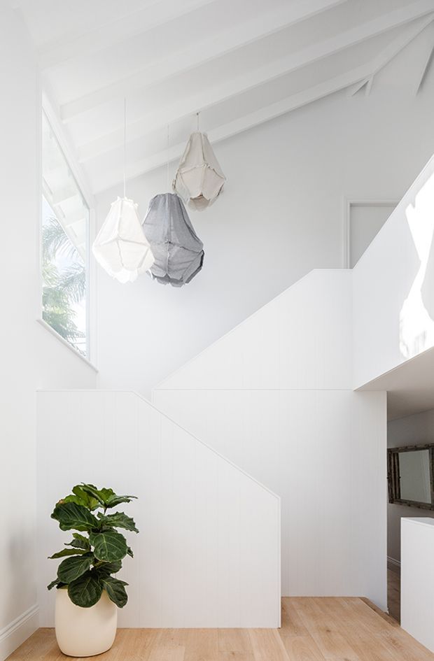A bright, predominantly white stairway with a sculptural geometric staircase, exposed white ceiling beams, and three hanging fabric pendant lights. Pale timber flooring, a potted leafy plant, and a partial mirror-lined room are visible, with daylight entering through tall glazing.