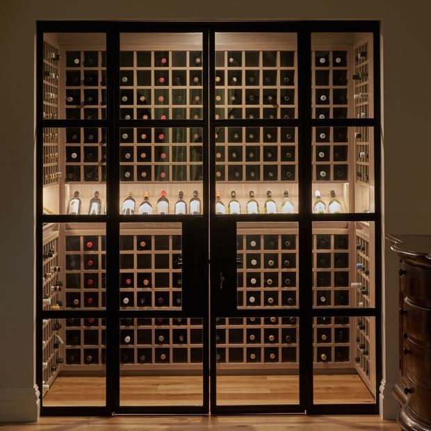 A dark-framed glazed double door opens onto a compact, floor-to-ceiling wine storage room. The room is lined with extensive timber wine racks and cubbies, filled with bottles, with illuminated display shelving and a warm timber floor. A dark traditional cabinet is partially visible at the right edge.