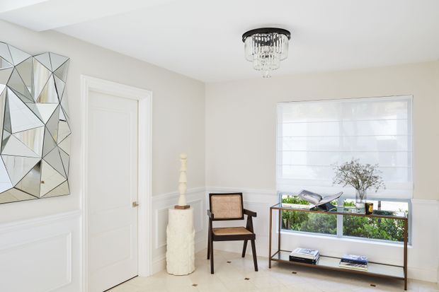 Bright, refined residential entry or hallway with white wall panelling, a closed white interior door, large geometric mirrored wall art, a crystal pendant light, and a slim dark-wood and glass console displaying decorative objects and books. A modern upholstered wood-framed chair and sculptural pedestal add understated decorative detail. The room has a light neutral palette and abundant daylight through a large window with a white Roman blind.