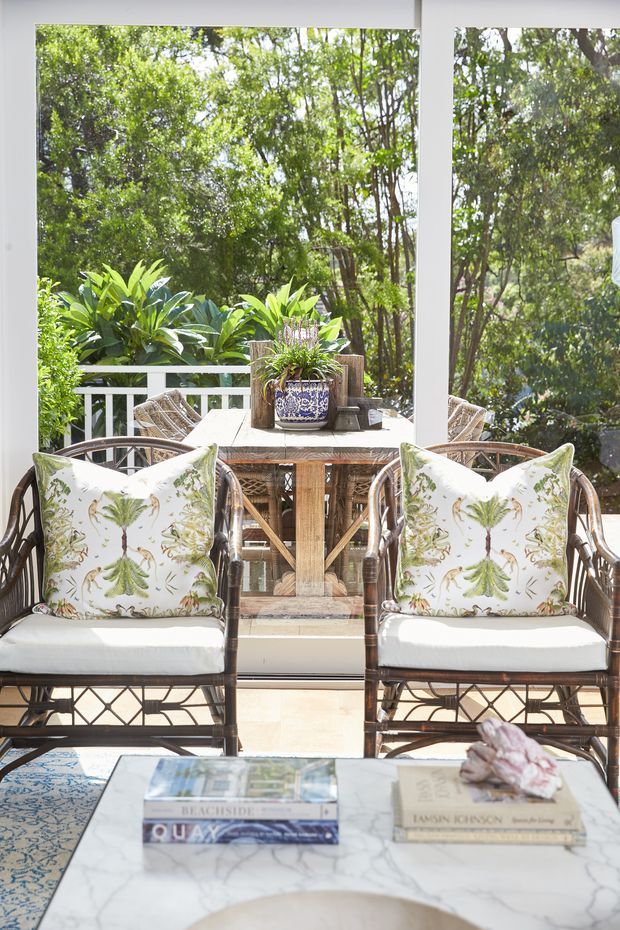 A bright, plant-filled sunroom or enclosed terrace with large white-framed windows overlooking dense greenery. Two rattan or wicker armchairs with white cushions and botanical-print pillows face a woven dining table and chairs, with a blue-and-white ceramic planter as a centrepiece. A marble-look coffee table, patterned rug, and stacked design books occupy the foreground. The space has an airy tropical, conservatory-like atmosphere.