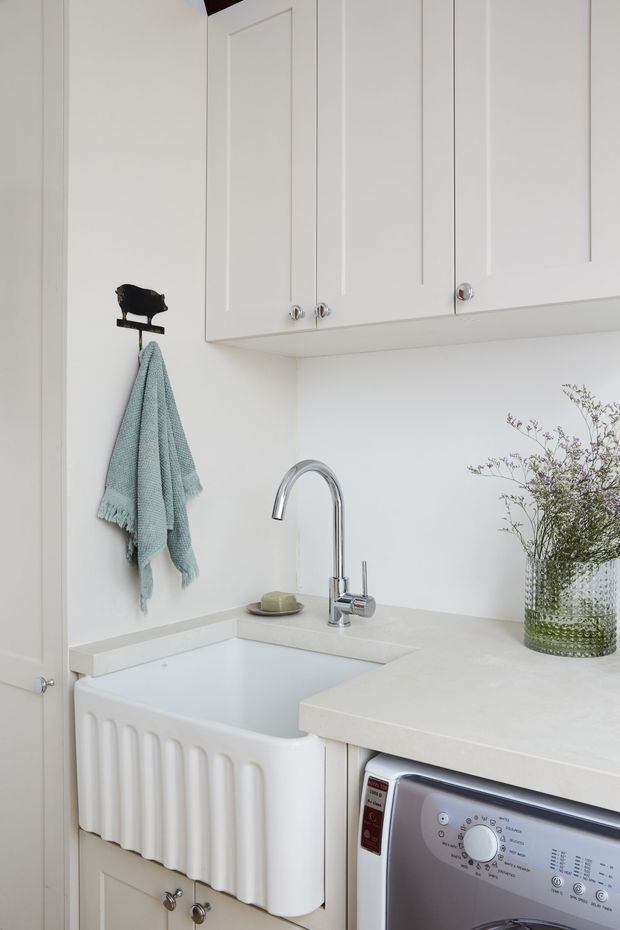 Bright, compact laundry room with white paneled overhead cabinetry, a deep white fluted farmhouse-style utility sink, pale benchtop, chrome gooseneck tap, and integrated front-loading washing machine. A pale blue towel and textured glass vase with delicate branches add restrained decorative detail.
