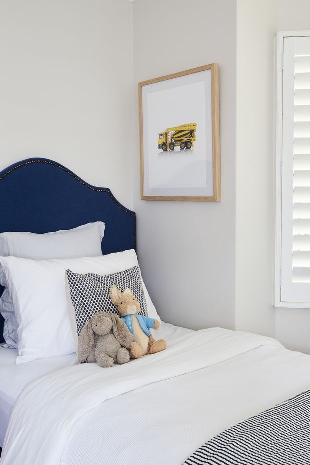 Bright, neatly styled children's bedroom featuring a single bed with crisp white bedding, a deep navy upholstered scalloped headboard with brass nailhead trim, plush toy animals, and a framed construction-vehicle print. Pale grey walls and white plantation shutters create a clean, cheerful scheme with subtle navy and patterned accents.