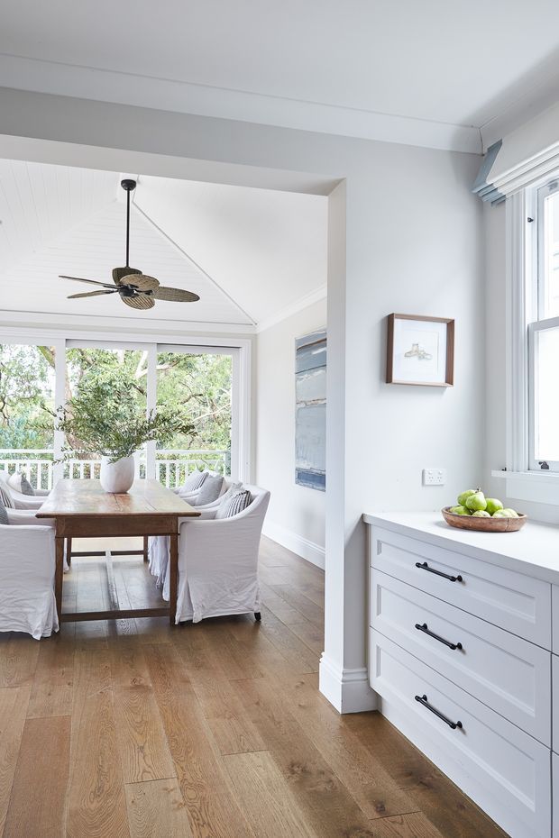 Bright, airy interior view looking from a built-in white storage area toward a dining room. The dining space has a rustic timber table, slipcovered upholstered chairs, a large leafy plant, and generous glazing opening toward a balcony or exterior greenery. White painted walls and ceiling, light timber flooring, white shaker-style drawers with dark hardware, framed artwork, and a suspended ceiling fan create a relaxed coastal-traditional feel.