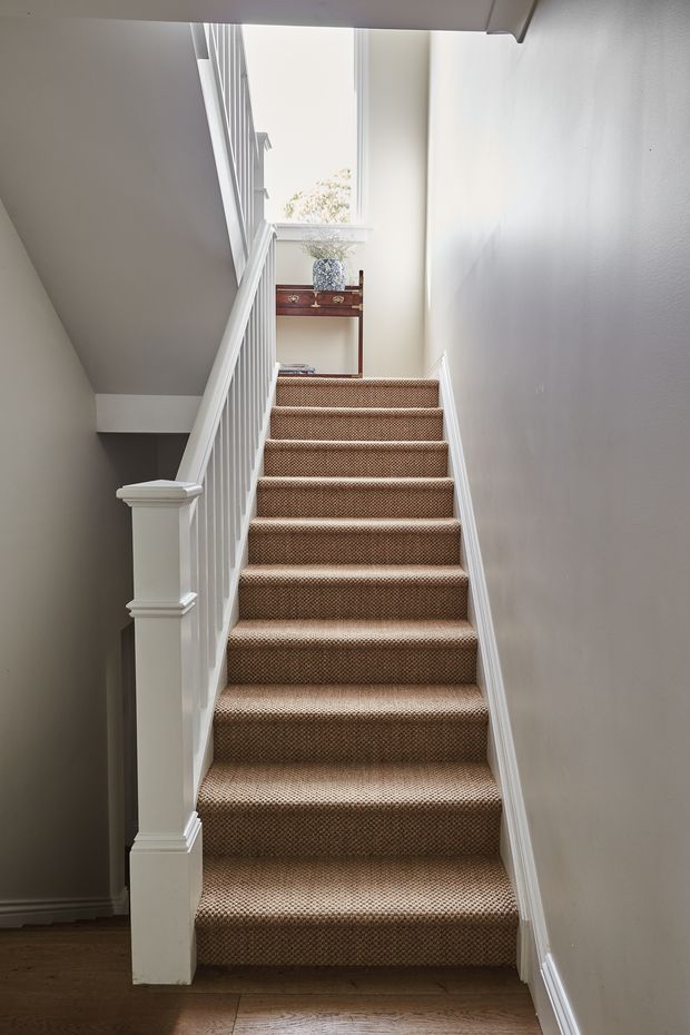 Interior photograph looking up a narrow residential staircase with brown patterned carpeted treads, white painted balustrades and trim, pale neutral walls, and timber flooring at the base. A small console table and decorative vase sit on the upper landing beside a bright window.