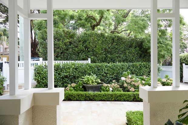 View from a covered residential entry or verandah through white square columns and open framing toward a manicured garden. The space has pale stone or tile paving, white painted architectural elements, low rendered walls, clipped hedges, flowering shrubs, and mature greenery beyond a white fence. The image is bright, calm, and strongly connected to the landscaped outdoor setting.