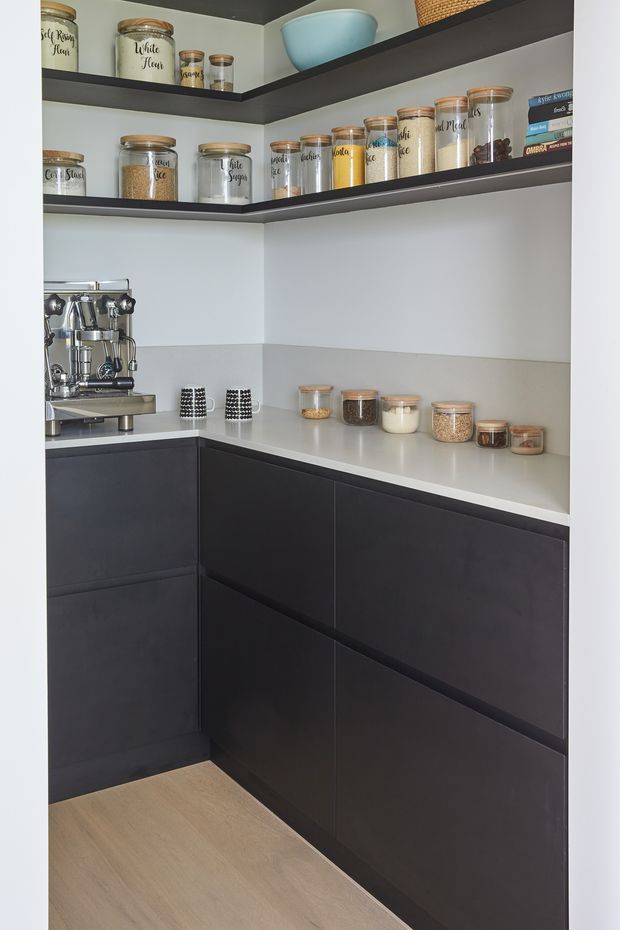 Compact, highly organised pantry with dark matte built-in cabinetry, white benchtops and backsplash, open dark shelving, and numerous labelled glass storage jars. A coffee machine and neatly arranged pantry staples are visible, with light timber flooring adding warmth.