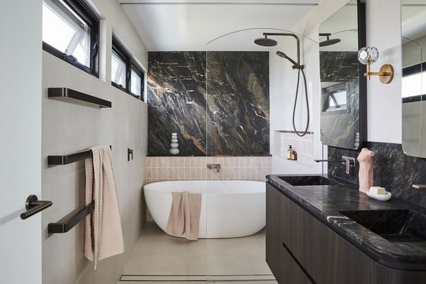A refined contemporary bathroom with a freestanding white bathtub, walk-in shower, dark veined stone feature wall and vanity benchtop, dark timber cabinetry, brass wall lighting, black tapware, and pale tiled flooring. Large mirrors and high horizontal windows add light and visual spaciousness.