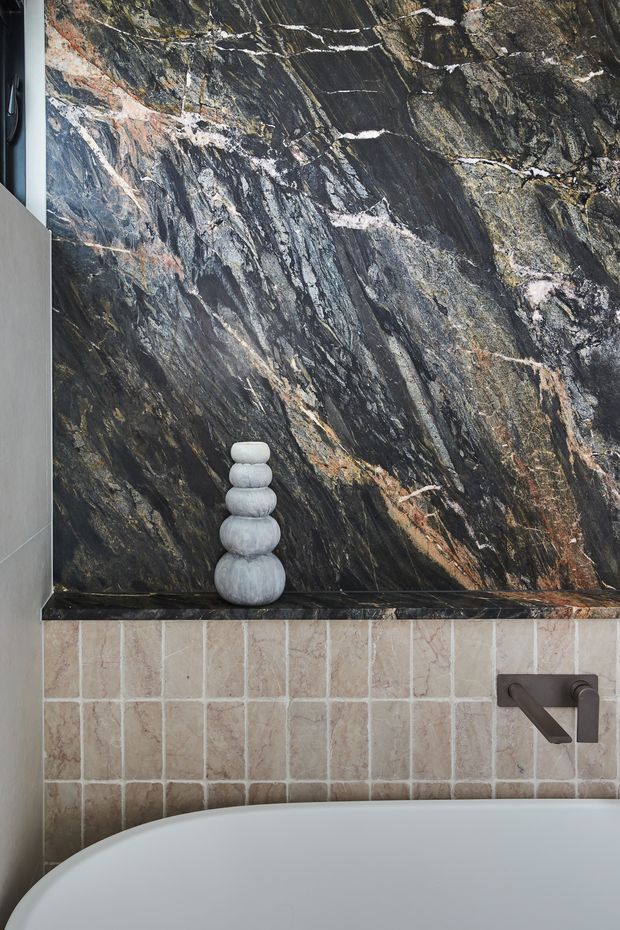 Close-up view of a refined bathroom feature wall. A dramatic dark veined natural stone slab forms the upper wall and recessed shelf, paired with vertically arranged pale beige stone tiles below. A freestanding white bath curves into the foreground, with a sculptural stacked-stone ornament and dark bronze wall-mounted tapware adding contrast.