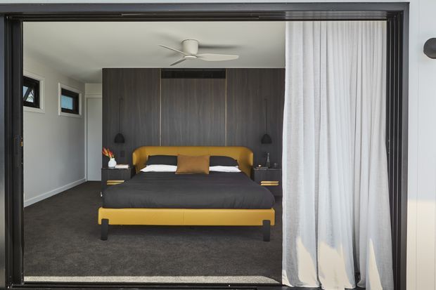 A contemporary bedroom viewed through a wide dark-framed opening, featuring a mustard yellow upholstered bed with dark bedding, a full-height dark timber feature wall and integrated bedside niches. The room has charcoal carpet, pale walls, small high-set windows, a ceiling fan, and a sheer white curtain partially drawn across the opening.