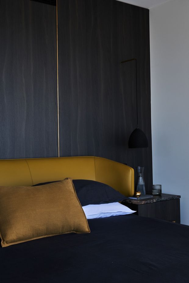A moody, refined bedroom vignette featuring a mustard-gold upholstered curved headboard, dark bedding, a large ochre cushion, and dark timber wall panelling. A small dark bedside table holds a glass bottle and tumbler, with a black pendant light suspended beside the bed. The palette is predominantly charcoal, black, deep wood, and ochre.