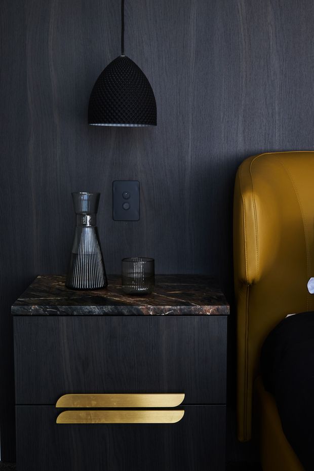 A moody, close-up interior detail showing a dark timber-look bedside cabinet with a heavily veined dark stone benchtop, oversized brushed-gold drawer pulls, and a black textured pendant light. Decorative glassware sits on the cabinet, while part of a mustard-gold upholstered bed or chair is visible at the right edge. The scene has a refined, dramatic, luxe aesthetic with strong dark-and-gold contrast.