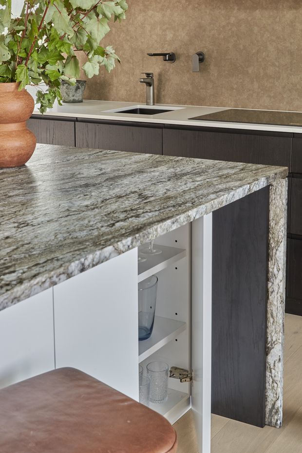 Close-up view of a contemporary kitchen featuring a large veined natural-stone island benchtop with an integrated open storage compartment, dark vertically textured cabinetry, a pale stone or composite rear benchtop, undermount sink, wall-mounted tapware and cooktop. Warm brown textured wall finish and a potted leafy plant add natural contrast.