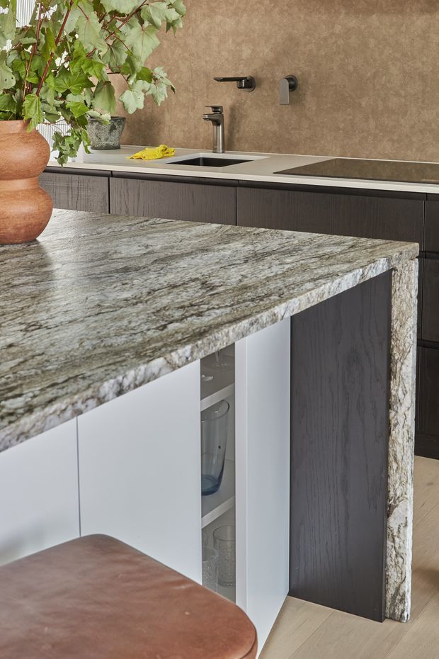 Close-up of a refined contemporary kitchen featuring a large natural stone island benchtop with a dramatic grey, white and taupe veining pattern. The island incorporates dark timber cabinetry, white storage fronts and an open display niche. In the background, matching dark cabinetry, an undermount sink, matte black tapware and an integrated cooktop sit against a warm textured brown backsplash. A leather-upholstered stool and potted greenery add warmth.