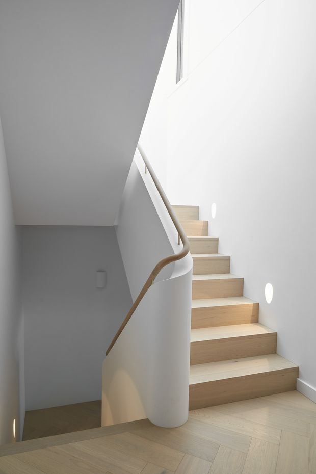 A minimalist interior stairway with pale timber treads and matching light timber flooring, enclosed by smooth white walls. The sculptural curved white balustrade features a rounded timber handrail, while small recessed wall lights illuminate the stairs. A high narrow window brings in natural light, creating a clean, bright, architectural composition.