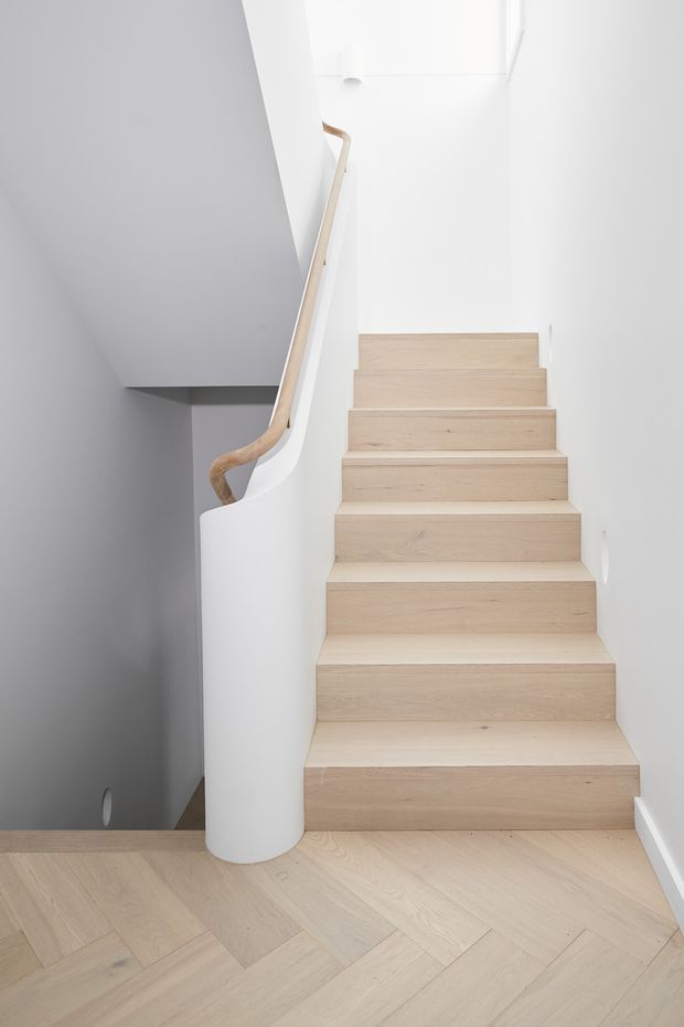 A minimalist interior stairway with pale timber treads and matching light timber herringbone flooring. The staircase has smooth white walls, a sculptural curved white balustrade, and a simple natural timber handrail. The space is bright, clean, and highly pared back.