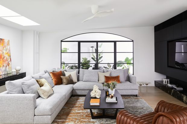 Bright contemporary living room with a large light-grey L-shaped sectional sofa, sculptural brown leather lounge chairs, a dark rectangular coffee table, and a textured neutral rug. A black-framed arched glass partition or conservatory-style window with indoor plants forms the backdrop, while a dark built-in media wall with television and low joinery occupies the right side. White walls, ceiling and ceiling fan create a clean, airy setting.
