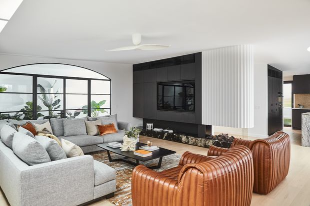 Bright contemporary living room with a large light-grey sectional sofa, two ribbed caramel-brown leather lounge chairs, a black rectangular coffee table, and a dark built-in media wall. A curved black-framed glazed partition opens to a plant-filled adjoining space, while a white fluted feature wall and pale timber flooring add texture and warmth. The room has a white ceiling with a modern ceiling fan and abundant natural light.