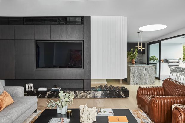 Contemporary open-plan living room with a dark built-in media wall, large television, sculptural curved white feature partition, stone fireplace hearth, grey sectional sofa, ribbed brown leather lounge chairs, and a black coffee table. The space opens toward a bright kitchen and dining area through large black-framed glazing.