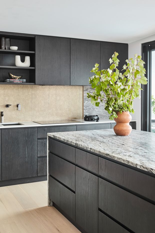 A refined contemporary kitchen with dark vertically grained timber cabinetry, integrated open shelving, a pale stone backsplash, and a large island topped with heavily veined natural stone. Light timber flooring, black-framed glazing, and a sculptural terracotta vessel with leafy branches add warmth and contrast.