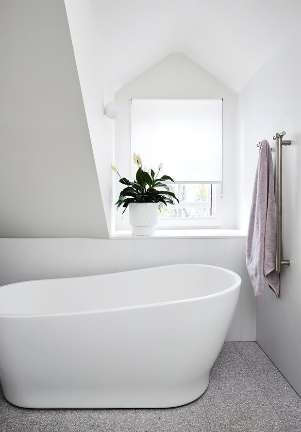 Bright minimalist bathroom with a freestanding white bathtub beneath a sloped ceiling and a large window fitted with a white roller blind. White tiled walls, a grey speckled floor, a potted plant on the window ledge, and a simple vertical towel rail with a mauve towel create a clean, serene interior.