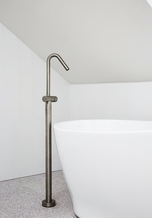 Minimalist bathroom corner featuring a freestanding white bathtub, tall brushed metal floor-mounted bath tap, white tiled walls, and a speckled terrazzo-style floor beneath a sloping ceiling.