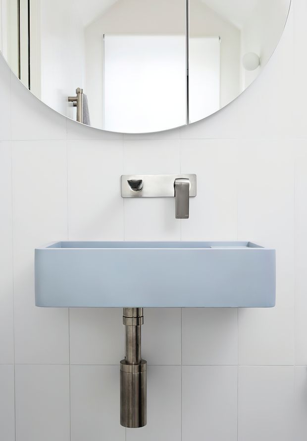 Minimalist bathroom vanity featuring a pale blue rectangular wall-mounted basin, brushed metal wall-mounted tap and exposed cylindrical bottle trap, set against white tiled walls. A large round mirror and simple wall light complete the clean, compact composition.