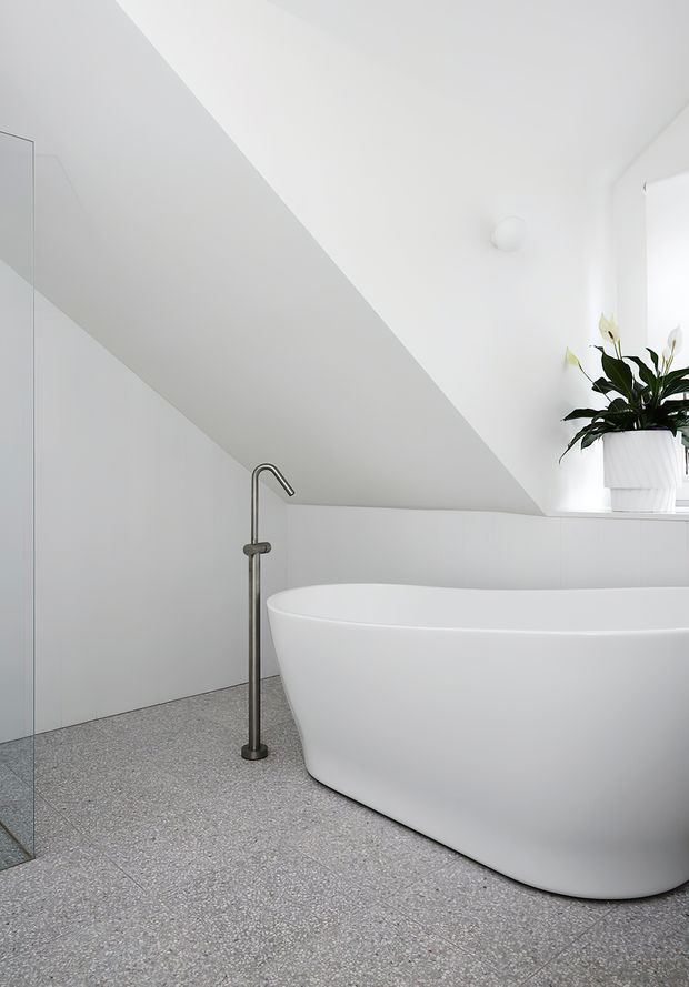 A bright minimalist bathroom beneath a sloping ceiling, featuring a freestanding white bathtub, wall-mounted or freestanding dark metal bath filler, pale grey terrazzo-style floor tiles, white wall surfaces, and a potted plant on a recessed ledge.