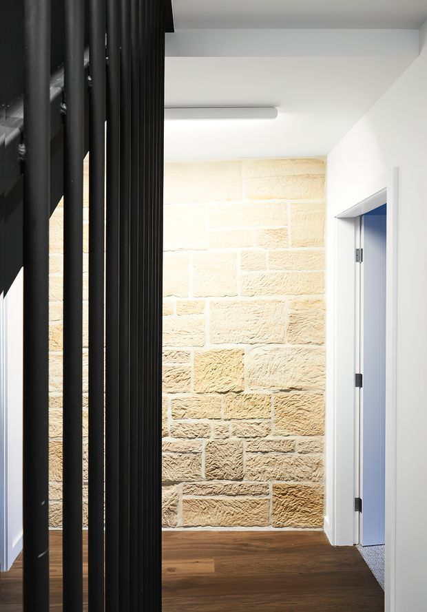 A bright modern hallway with a warm textured sandstone block feature wall, dark timber flooring, white walls and ceiling, and a black vertical slatted screen along the left side. A white-framed doorway opens on the right, while a linear ceiling light provides illumination.