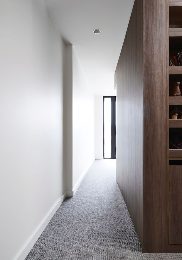 A narrow, bright residential hallway with white painted walls, a recessed ceiling spotlight, grey textured carpet, and dark timber built-in shelving and cabinetry along the right wall. A slim glazed door or window at the far end provides daylight. The space is clean and minimalist with strong linear perspective.