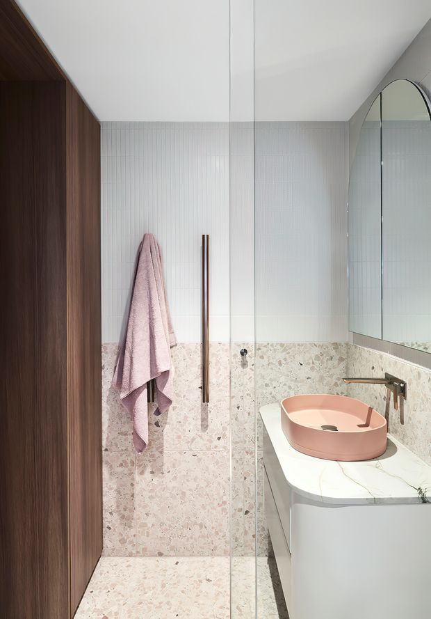 A compact contemporary bathroom with a walk-in shower, pale terrazzo wall and floor tiles, white vertical wall tiling, a curved white vanity with a pale pink vessel basin, brushed metal wall-mounted tapware, dark timber joinery, and a large arched mirror.
