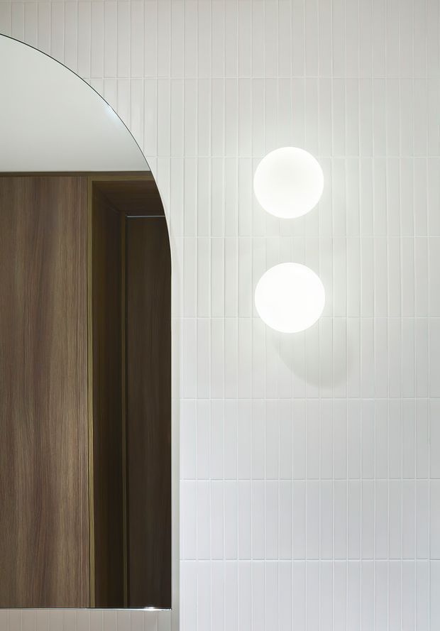 Close-up view of a refined bathroom wall featuring vertically stacked narrow white tiles, two round globe wall lights, and a large arched mirror with a dark timber-framed or timber-lined vanity area reflected within it. The composition is minimalist and carefully detailed, with warm wood contrasting against the bright tiled surface.