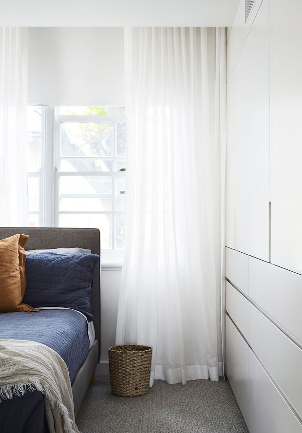 Bright, compact bedroom corner with a low upholstered bed dressed in navy blue bedding and layered throws, a woven basket on a grey carpet, sheer white curtains over a window, and full-height white built-in cabinetry with sleek integrated drawer fronts. The space has a clean, light-filled contemporary appearance.