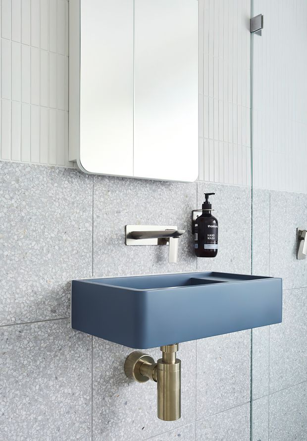 A compact contemporary bathroom vanity with a deep blue wall-mounted basin, brushed brass exposed waste pipe and wall-mounted tap, terrazzo-style grey wall tiles, and white vertically lined wall tiles. A rounded-corner mirrored cabinet hangs above the basin, with a black handwash dispenser and partial shower hardware visible at right.