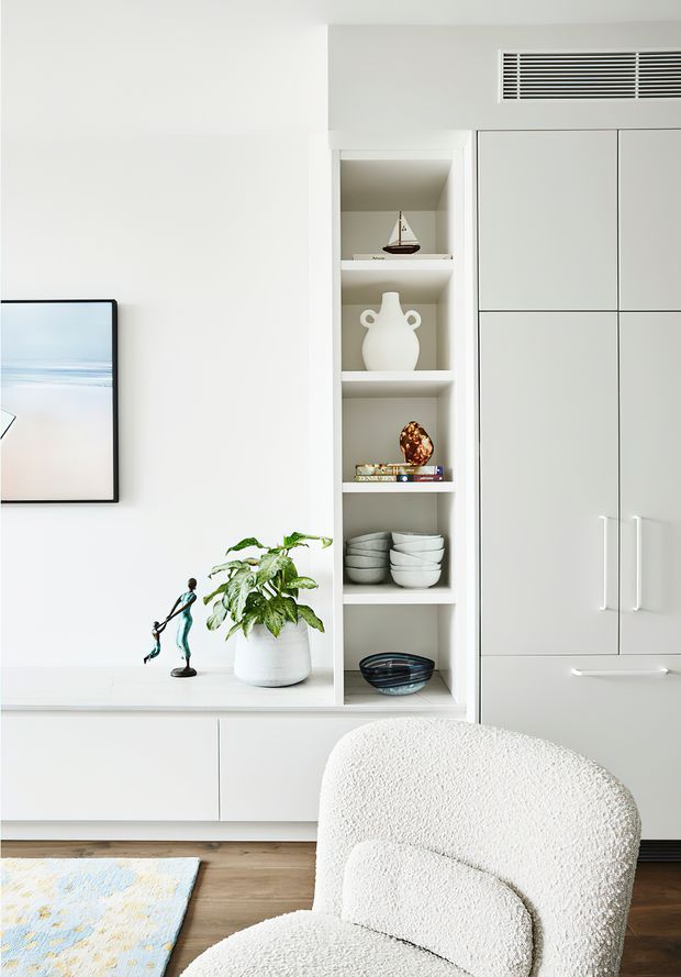 Bright, minimalist living room detail featuring a white built-in entertainment and storage wall with open display shelving, concealed cabinetry, decorative objects, and a wall-mounted television. A textured boucle armchair sits in the foreground, with a potted plant, light timber flooring, and pale rug adding warmth.