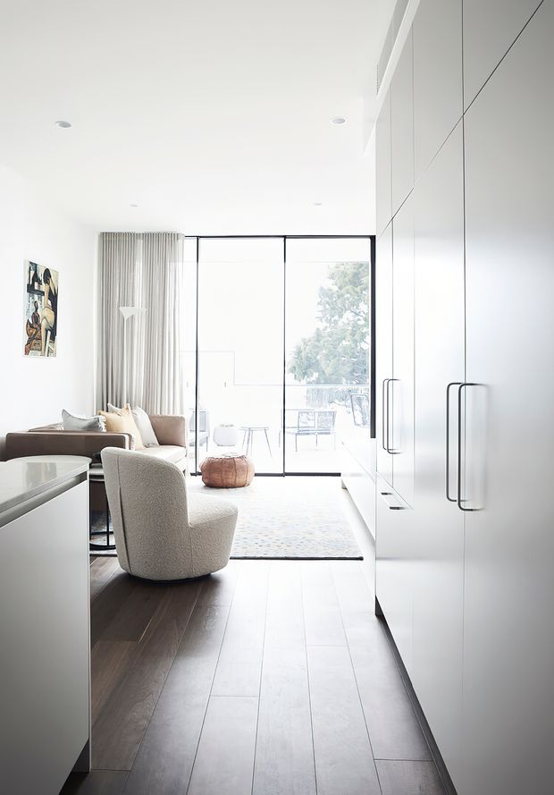 Bright, refined open-plan interior viewed from a narrow circulation area, with full-height white built-in cabinetry and long dark handles on the right, dark timber flooring, a neutral seating area, soft curtains, and floor-to-ceiling glazed doors opening to an outdoor balcony or terrace. The space has a clean, contemporary minimalist character with abundant natural light.