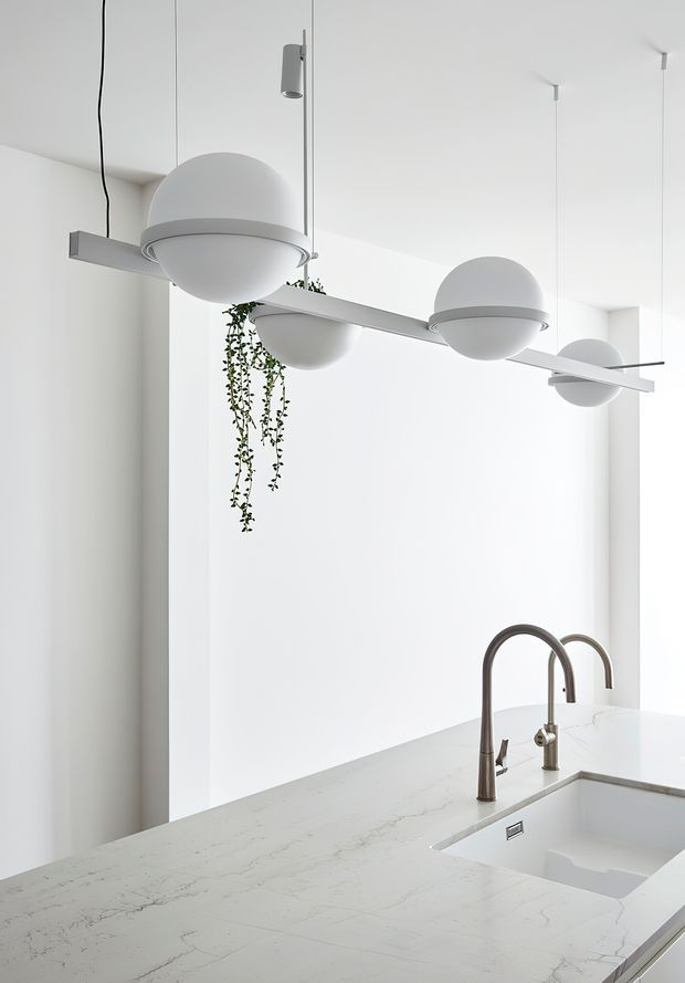 Bright minimalist kitchen interior featuring a large white stone island with integrated sink, tall curved brushed-metal tapware, and sculptural suspended globe-and-bar pendant lights. White walls, high ceilings, and sparse hanging greenery create a clean, airy composition.