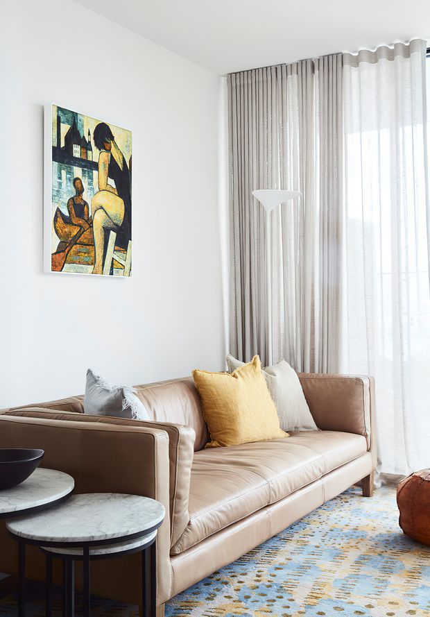 A bright, refined living room featuring a blush-beige leather sofa with yellow, cream and grey cushions, layered round marble-top nesting tables, a sculptural white floor lamp, patterned blue-and-gold rug, abstract figurative artwork, and full-height sheer and textured curtains at a large window.