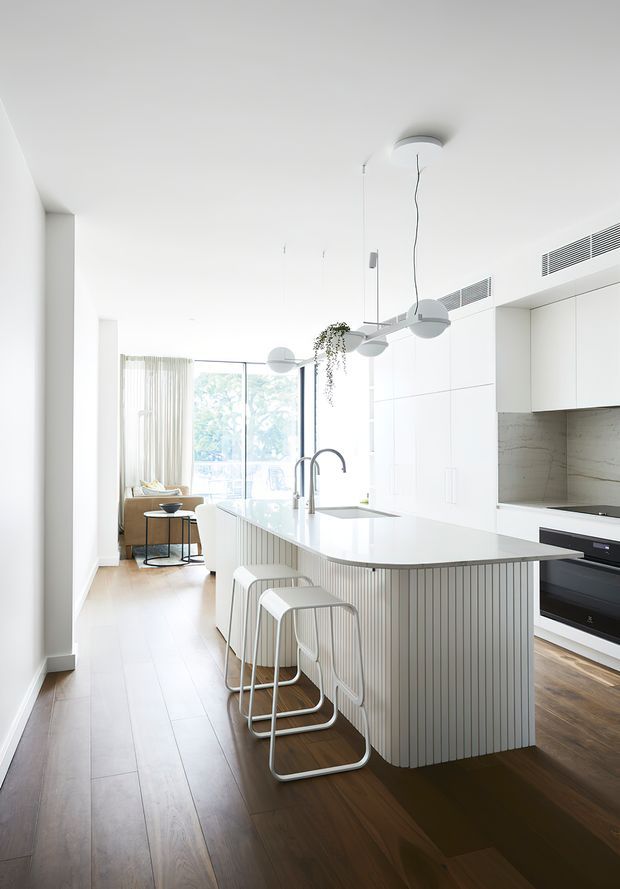 Bright contemporary open-plan kitchen with a large white island featuring vertical fluted detailing, integrated sink and curved white bar stools. White full-height cabinetry, a pale stone splashback, timber flooring, sculptural pendant lights, and floor-to-ceiling glazing create an airy minimalist interior. A small living area is visible beyond the kitchen.