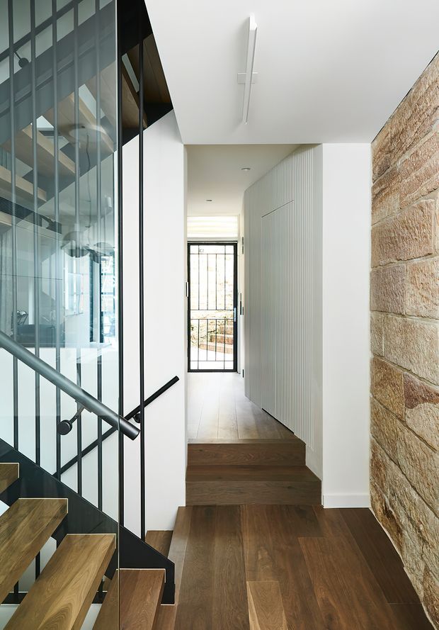 A bright contemporary hallway and stair landing with dark timber flooring and stairs, a black-framed glazed stair balustrade, white walls and ceiling, a vertical-ribbed white built-in wall, and an exposed warm sandstone feature wall. A glazed door at the end opens toward an exterior area. The space combines crisp modern detailing with natural timber and masonry textures.