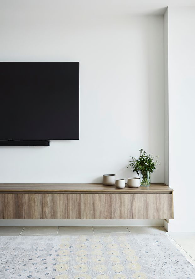 A minimalist living room media wall with a wall-mounted black television above a long floating timber-look entertainment cabinet. The cabinet has clean-lined doors and a matching wood-grain top, styled with neutral ceramic vessels and a glass vase of greenery. White walls, pale tiled flooring, and a muted patterned rug create a bright, restrained interior.