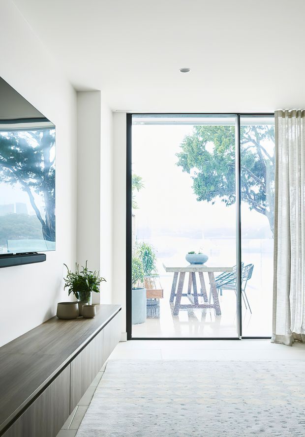 Bright contemporary living room with a minimalist built-in media console, wall-mounted television, indoor plants, pale rug, and full-height glazed sliding doors opening to a terrace with outdoor furniture and water views beyond. The room has white walls and ceiling, dark slim window frames, and sheer neutral curtains.